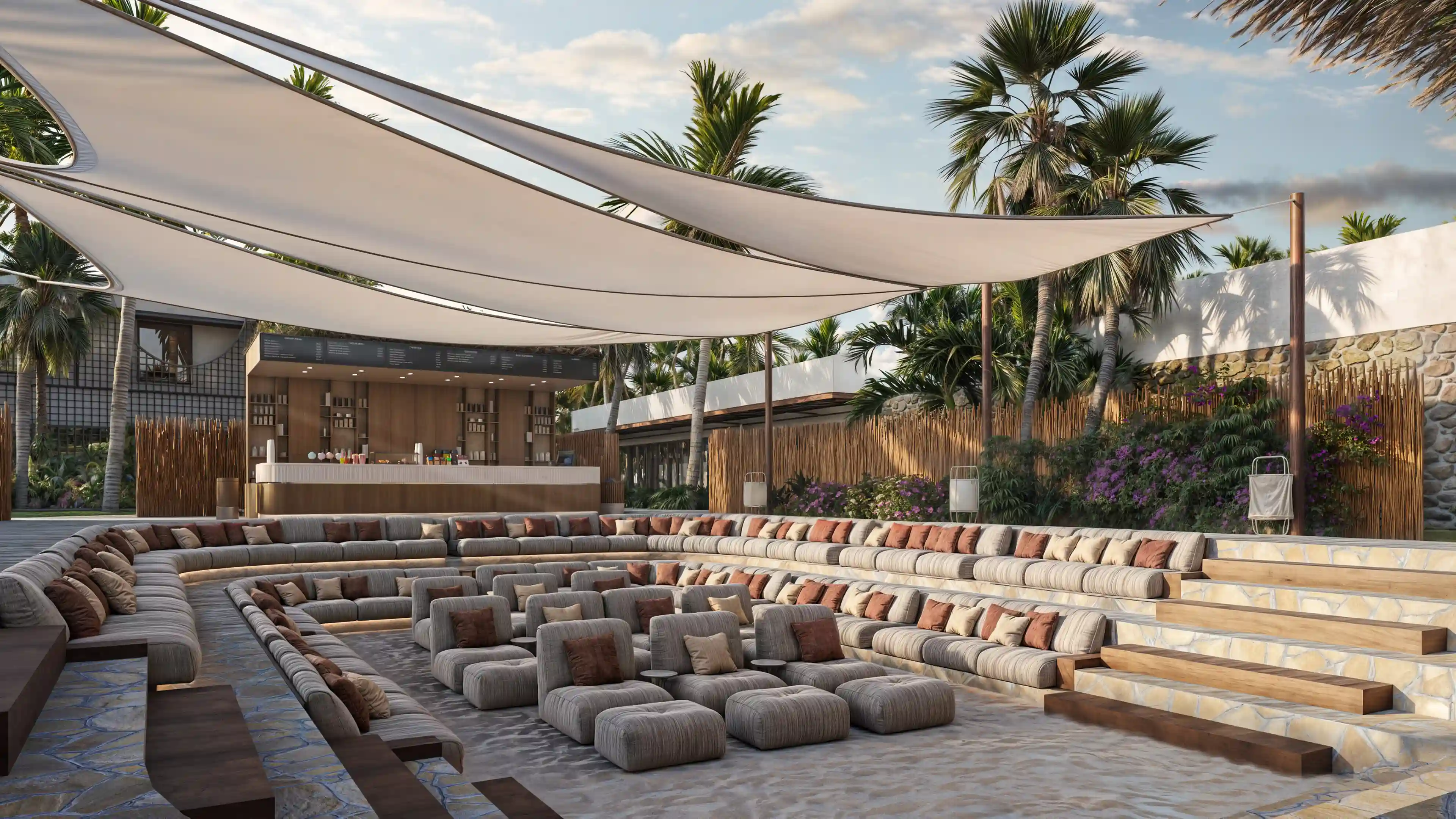 Outdoor sunken seating area with cushioned sofas and chairs under white shade sails, surrounded by palm trees and a bar counter in the background.