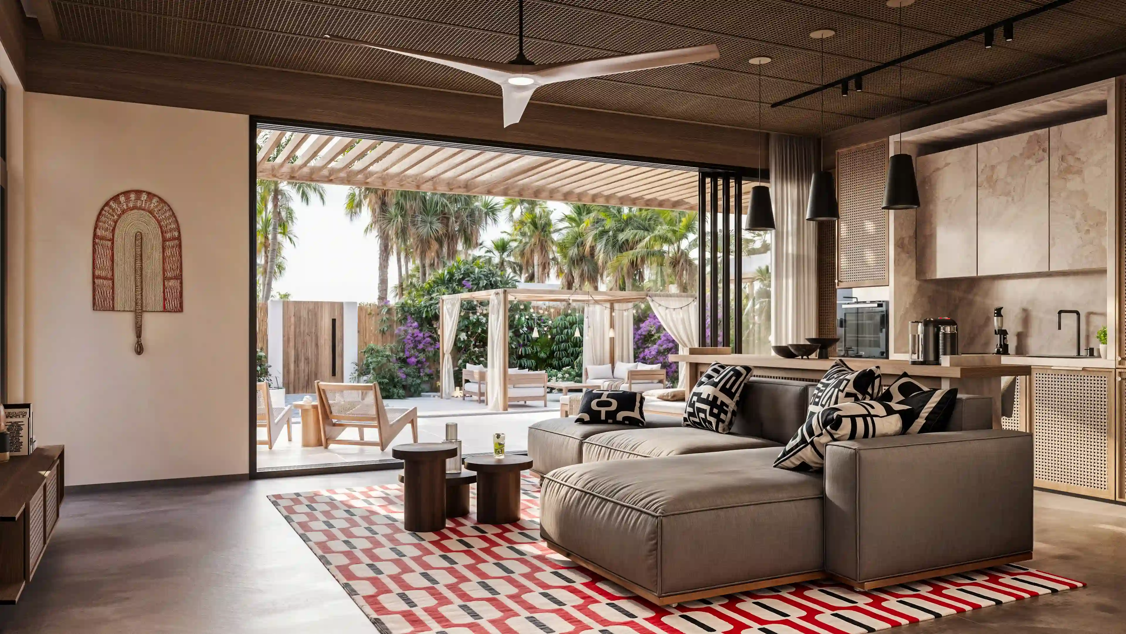 Modern living room with a gray sectional sofa adorned with black and white patterned pillows, two round wooden coffee tables on a geometric rug, and an open view to an outdoor patio with palm trees and shaded seating.