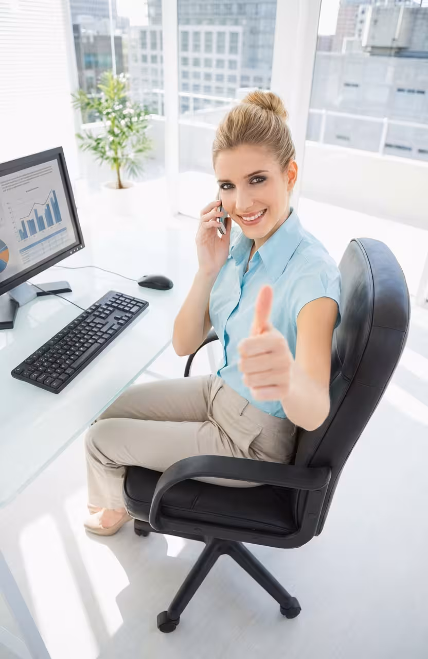 Smiling woman at office desk talking on phone and giving thumbs up, with a computer monitor displaying charts nearby.