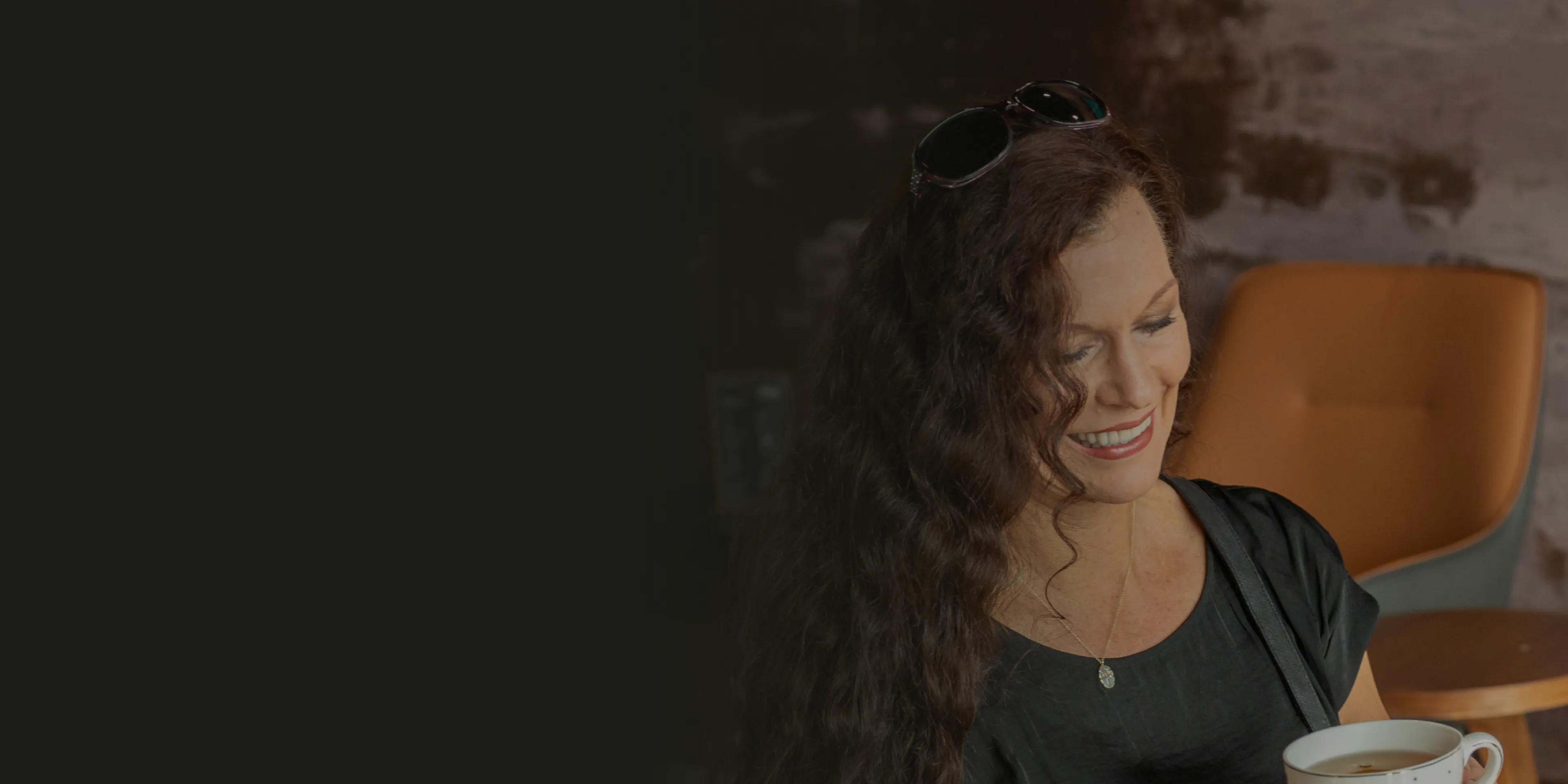 A woman with curly hair smiles while holding a white cup against a dark background.