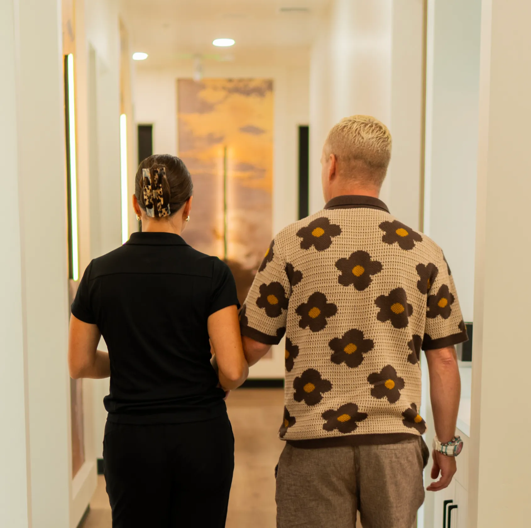 Two people walking down a hallway, one guiding the other by the arm.
