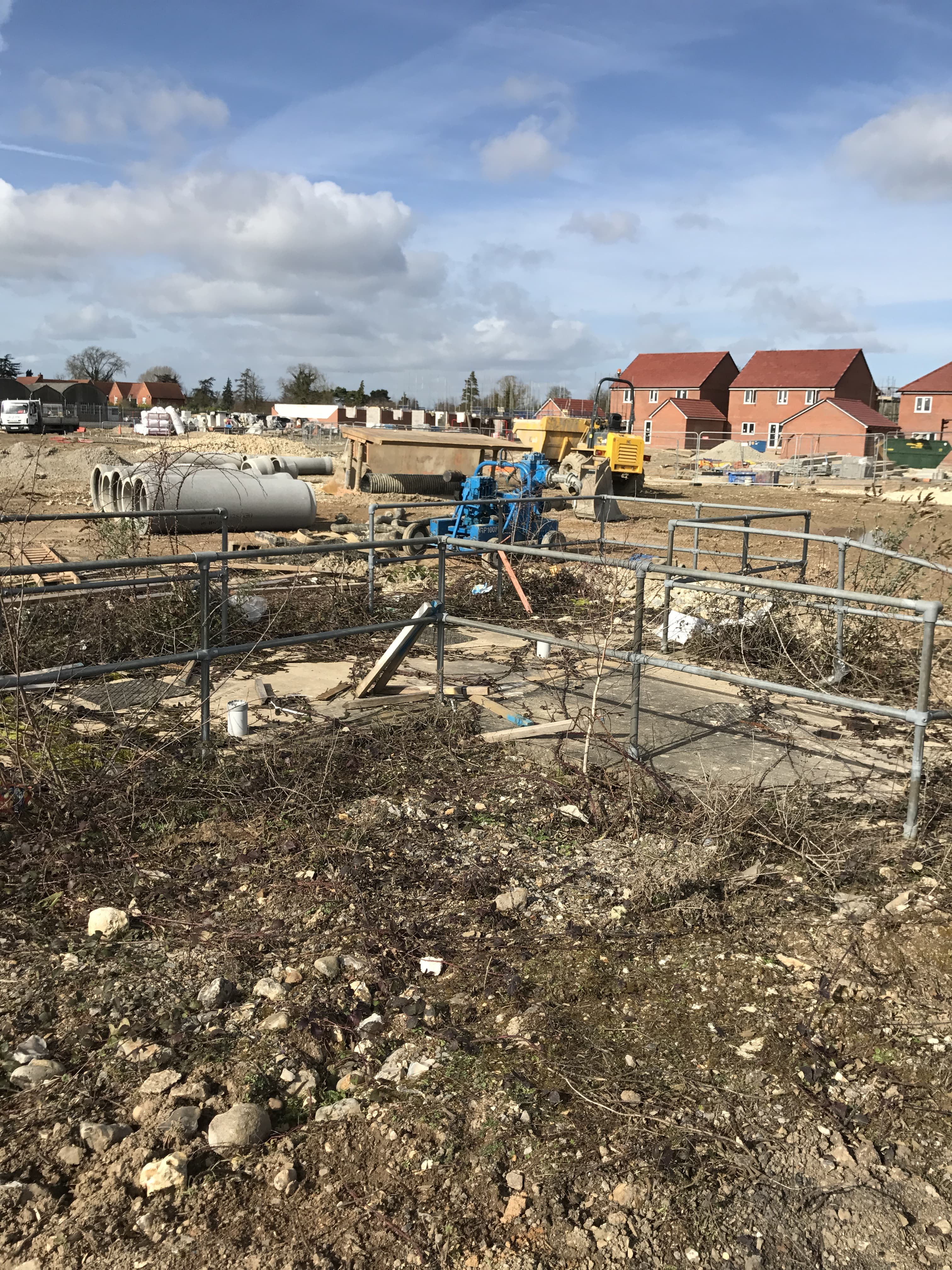 Construction site with pipes, equipment, and fenced-off areas, with newly built houses in the background under a partly cloudy sky.