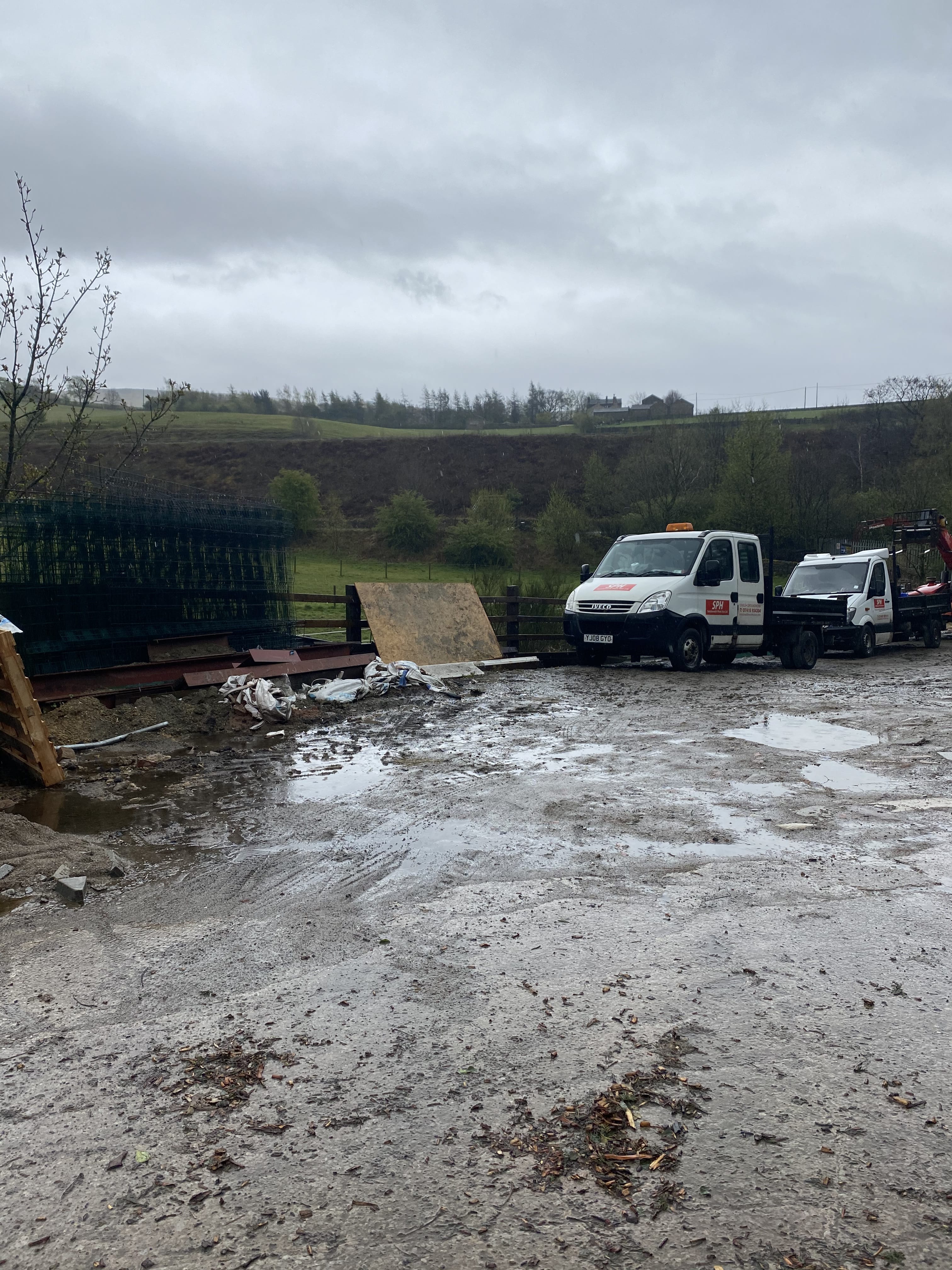 Mud-covered construction site with two small white work trucks parked near fencing materials under a gray cloudy sky.