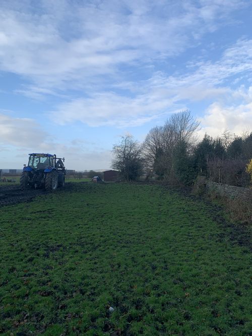 Blue tractor driving on a muddy green field next to leafless trees under a partly cloudy sky.