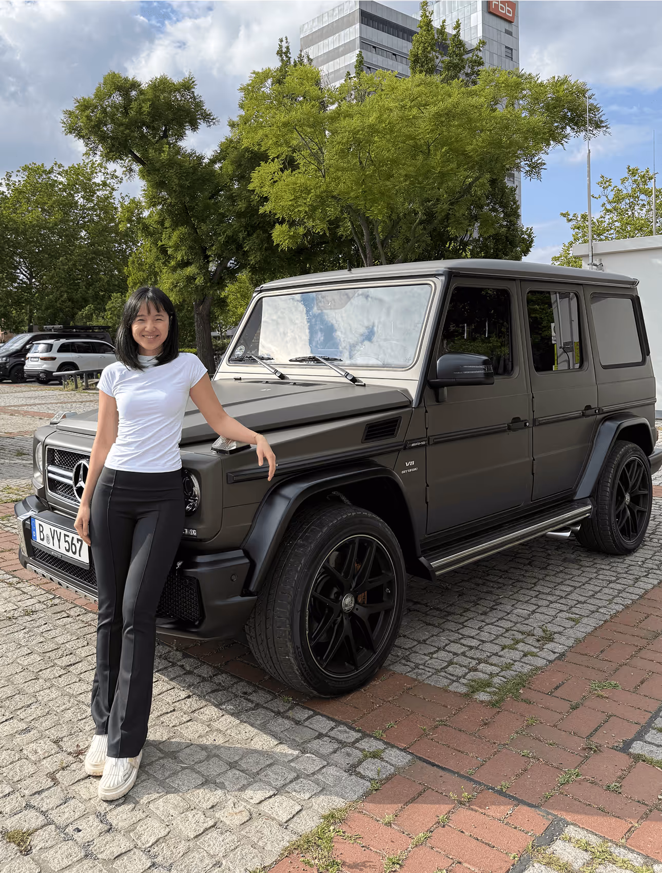 Frau neben schwarzem Mercedes-Geländewagen auf gepflastertem Platz, umgeben von Bäumen und modernen Gebäuden.