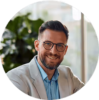 Ein lächelnder Mann mit Brille und Bart, trägt einen hellen Anzug und blauen Hemd in einem sonnigen Büro mit Pflanzen im Hintergrund.
