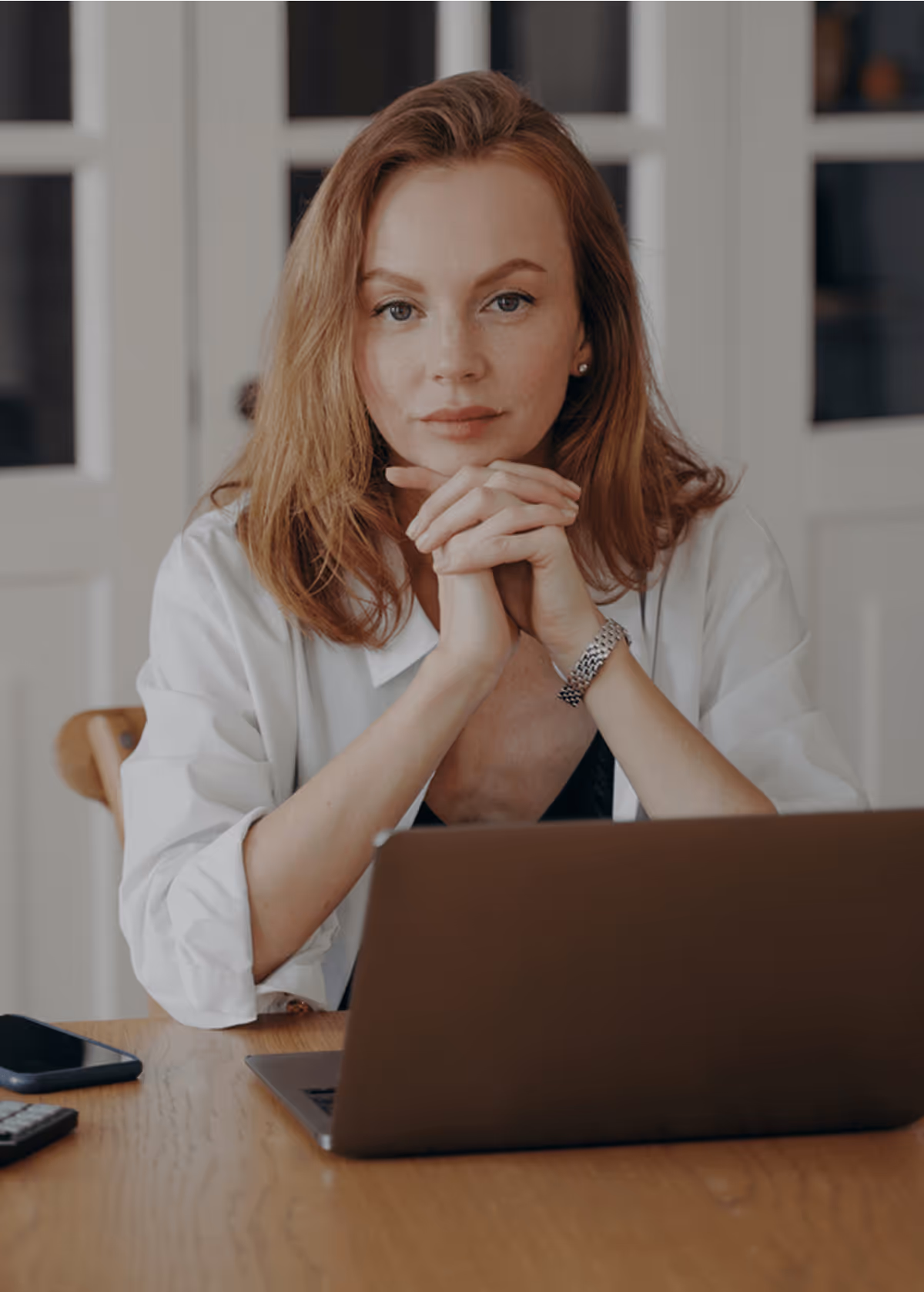 Frau mit Laptop am Tisch, konzentrierter Blick, weiße Bluse, Smartphone daneben, moderne Arbeitsumgebung.