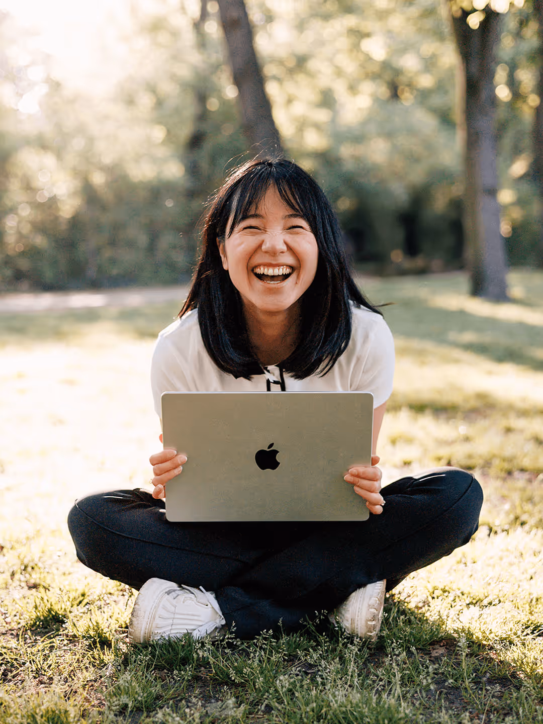 Frau sitzt lächelnd im Park, hält Laptop mit Apple-Logo, sonniger Tag, umgeben von Bäumen.