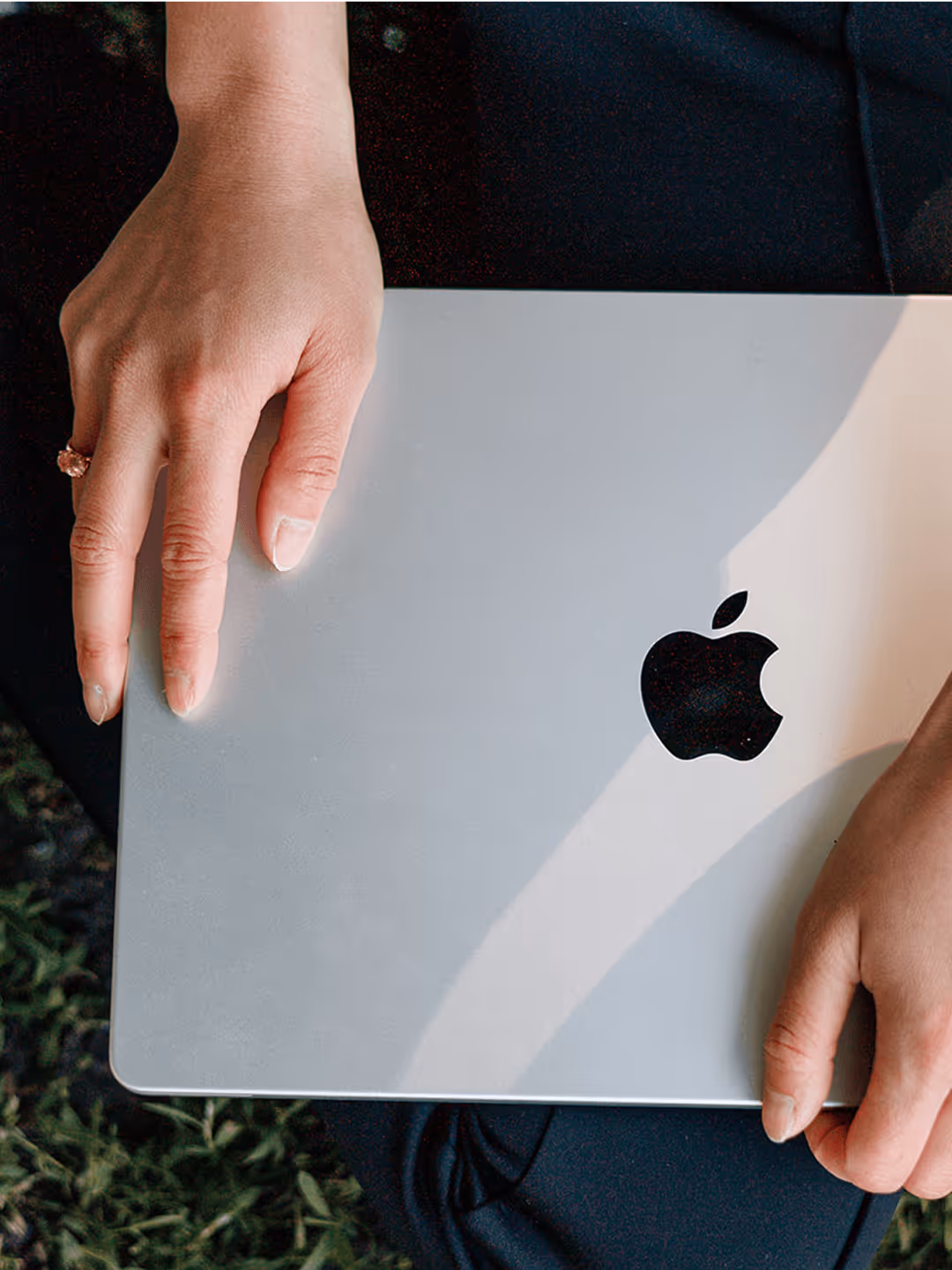 Person hält MacBook, auf dem Schoß liegend, mit Apfel-Logo sichtbar. Hände zart auf dem Laptop platziert, Gras im Hintergrund.