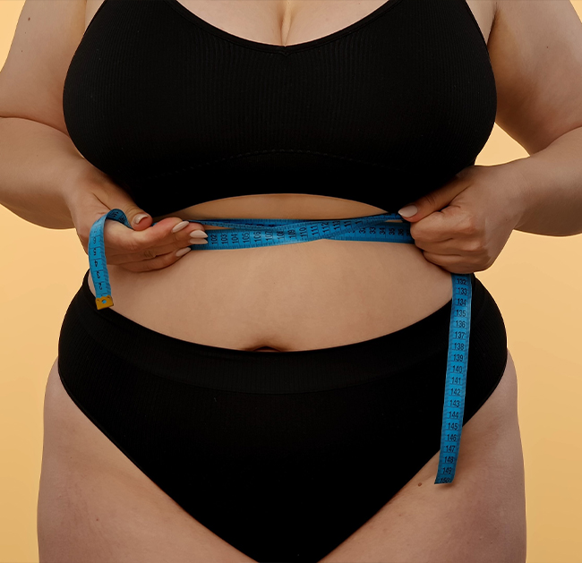 Person in black underwear measuring waist with a blue measuring tape against a beige background.