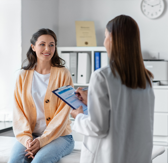 Smiling woman in a peach cardigan sitting on an examination table while a doctor in a white coat holds a clipboard and pen.