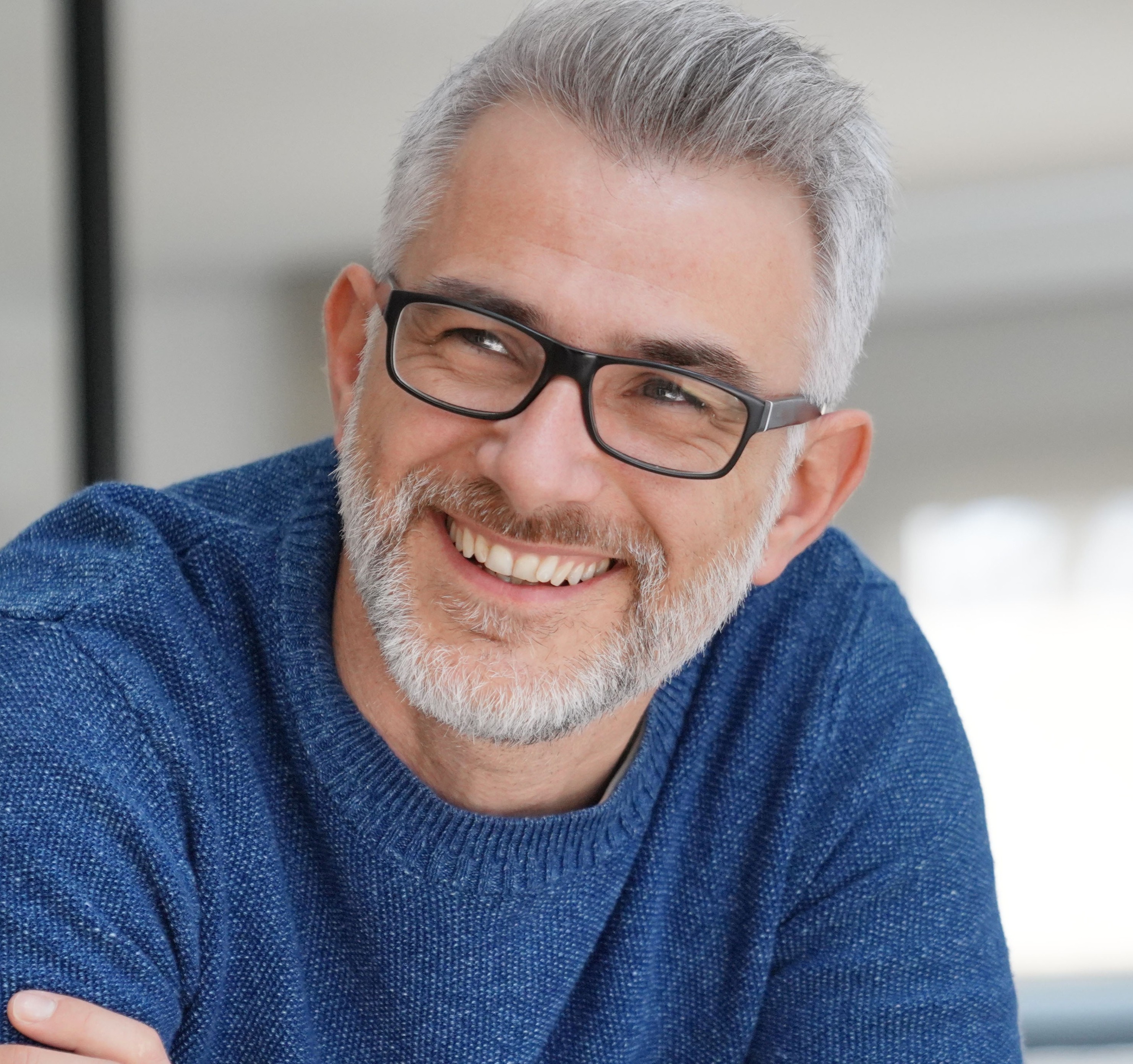 Smiling middle-aged man with gray hair, beard, black glasses, and a blue sweater looking to the side.