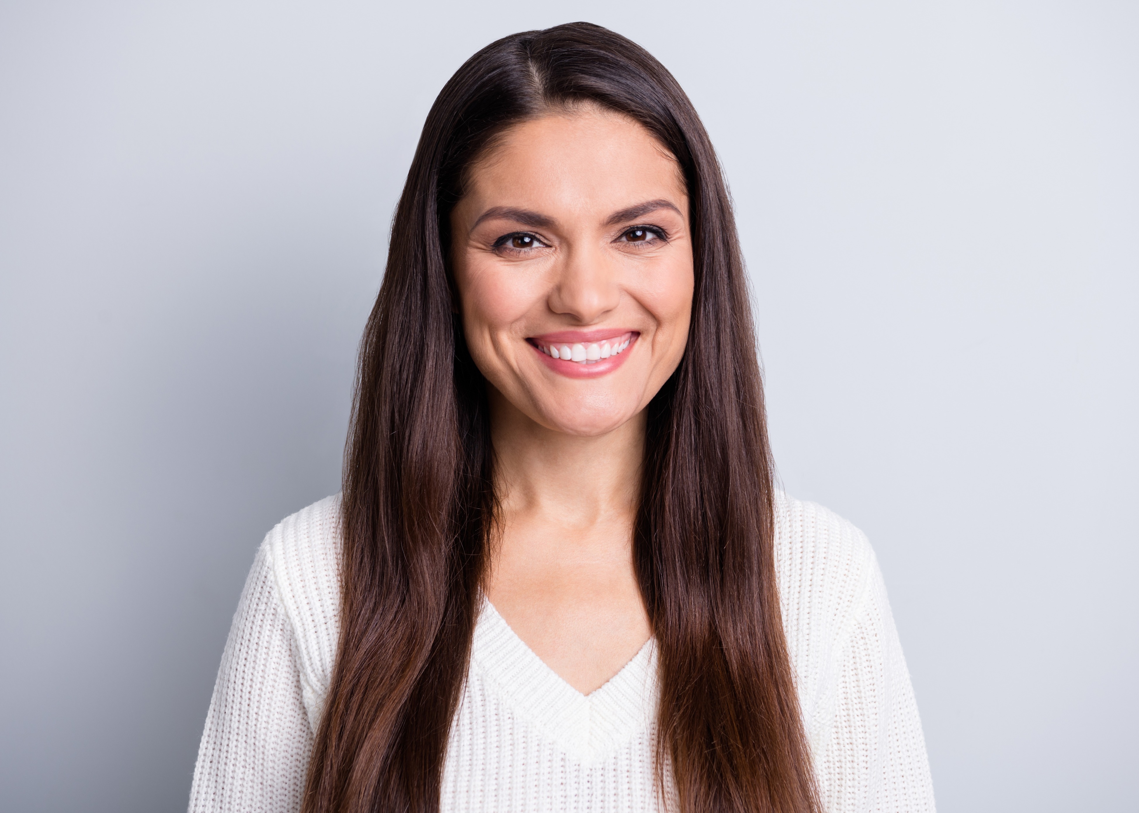 Smiling woman with long dark hair wearing a white knit sweater against a gray background.