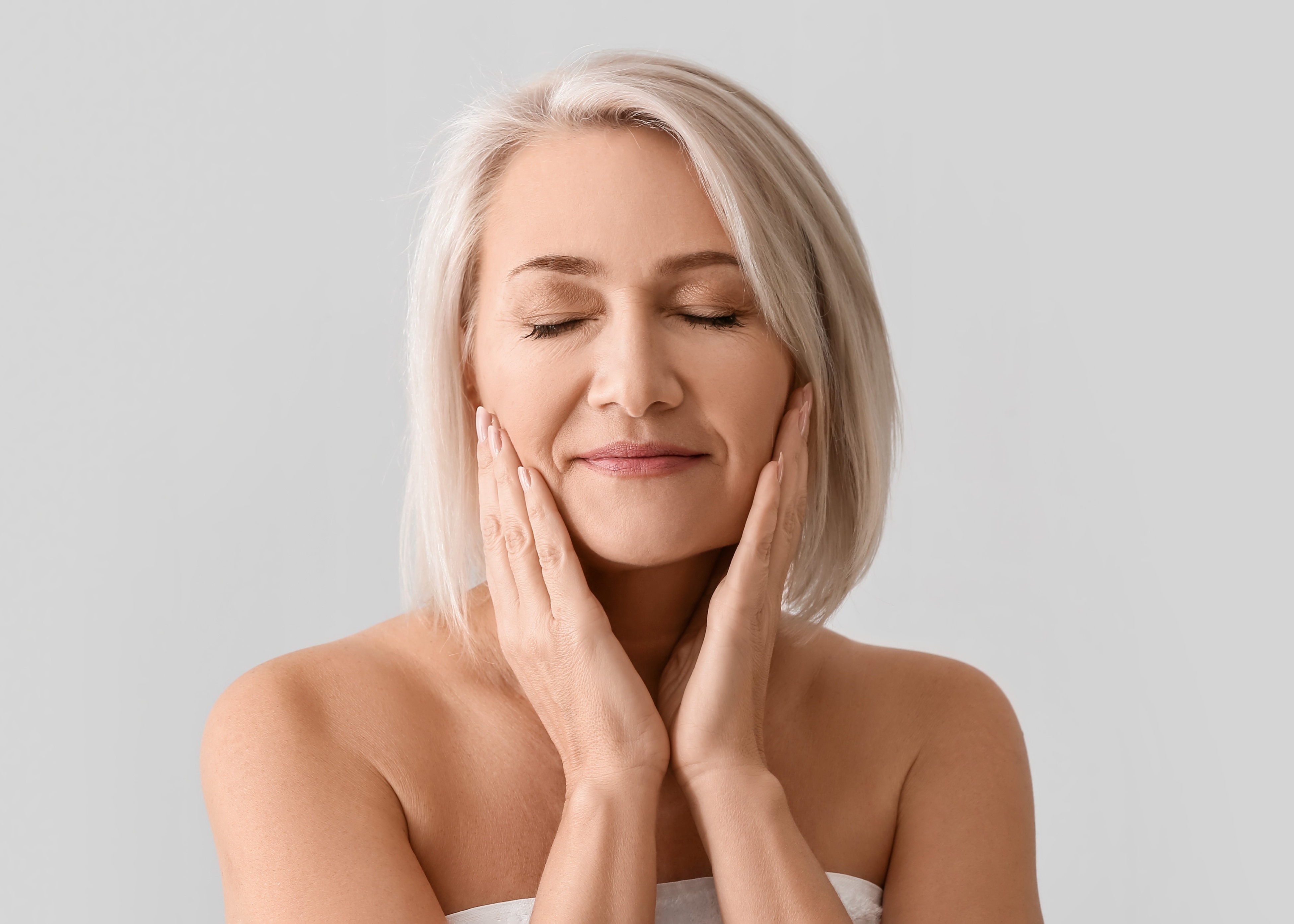 Middle-aged woman with closed eyes gently touching her face with both hands against a plain gray background.