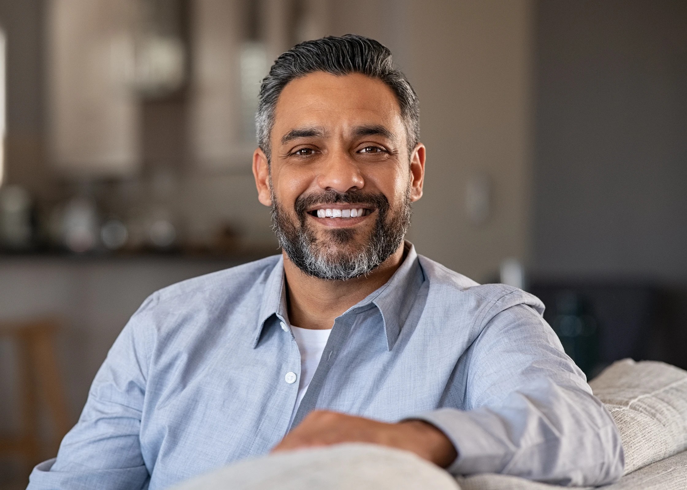 Smiling middle-aged man with salt-and-pepper hair and beard wearing a light blue shirt sitting on a couch indoors.