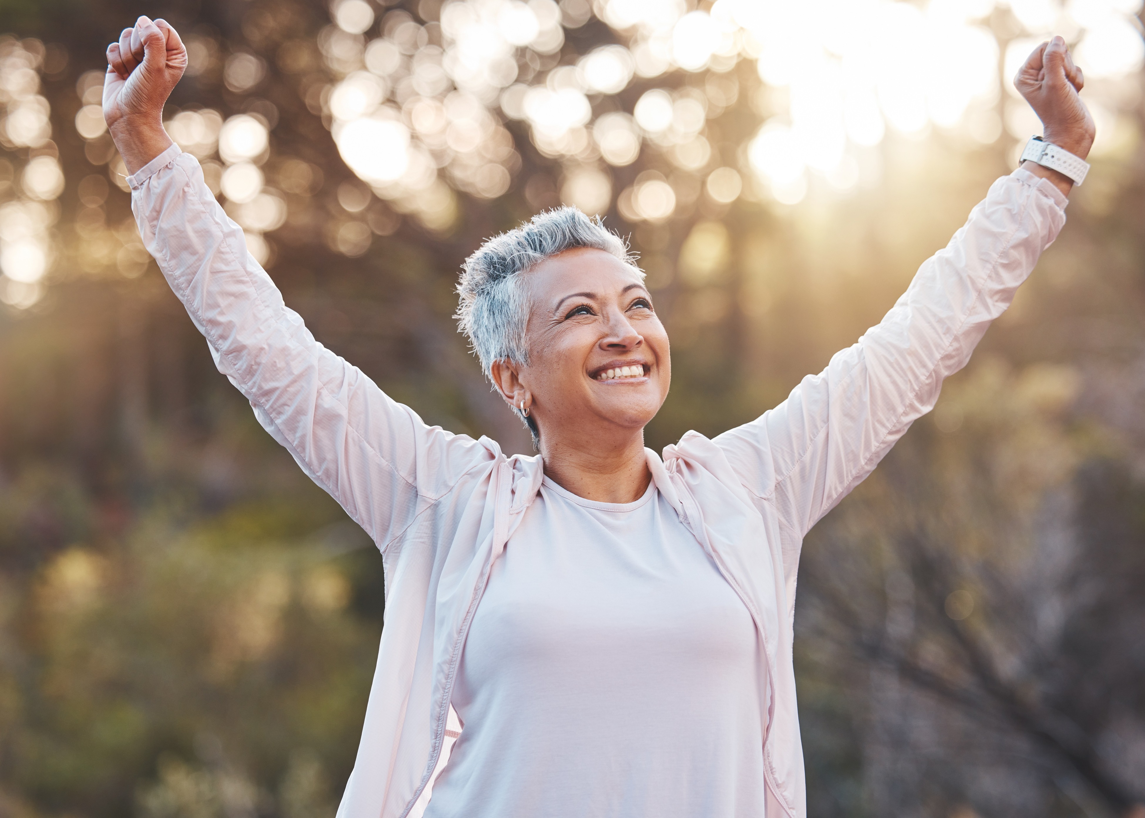 Smiling middle-aged woman with short gray hair raising her arms outdoors during golden hour.