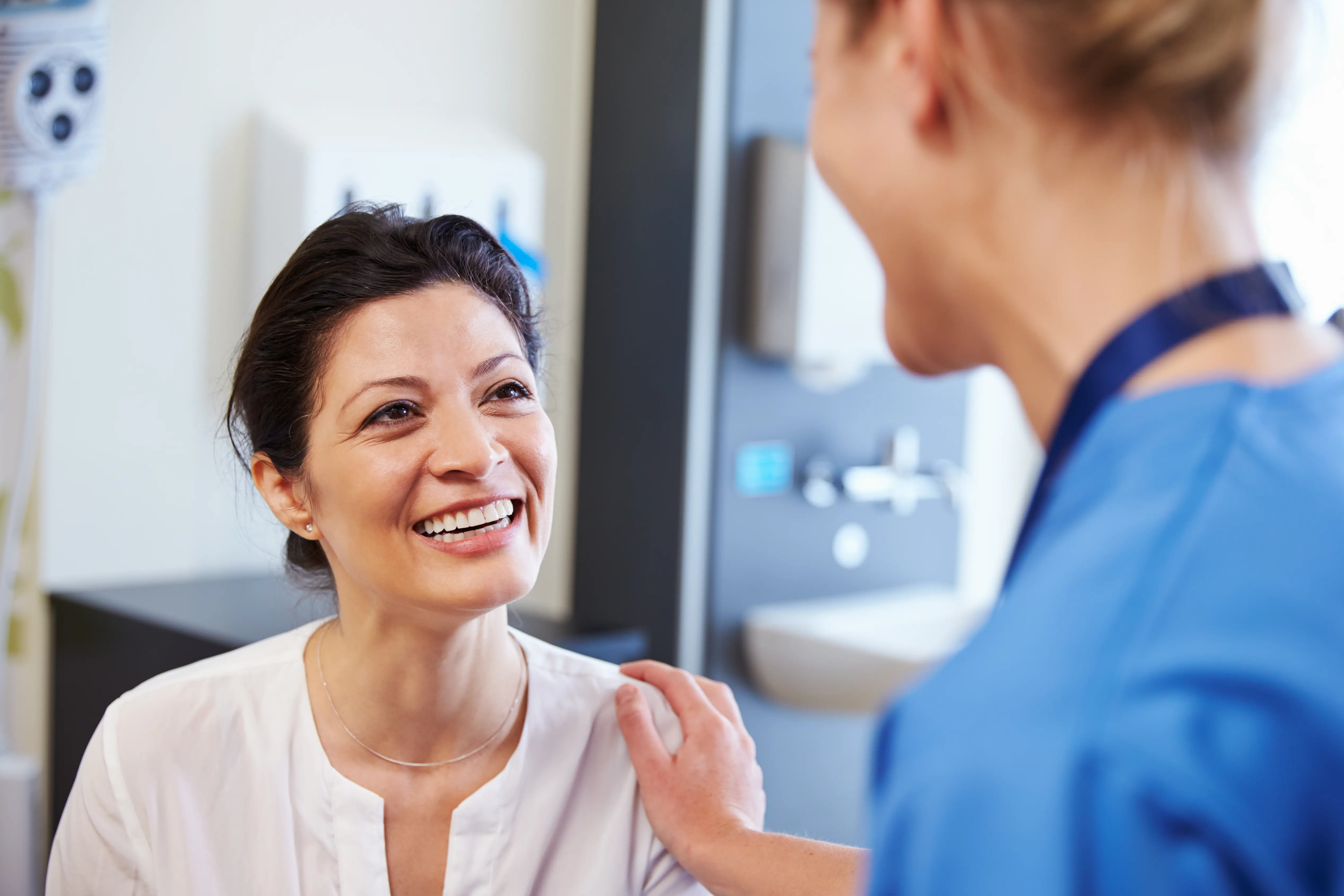 Smiling woman in white shirt being comforted by a healthcare worker in blue scrubs.