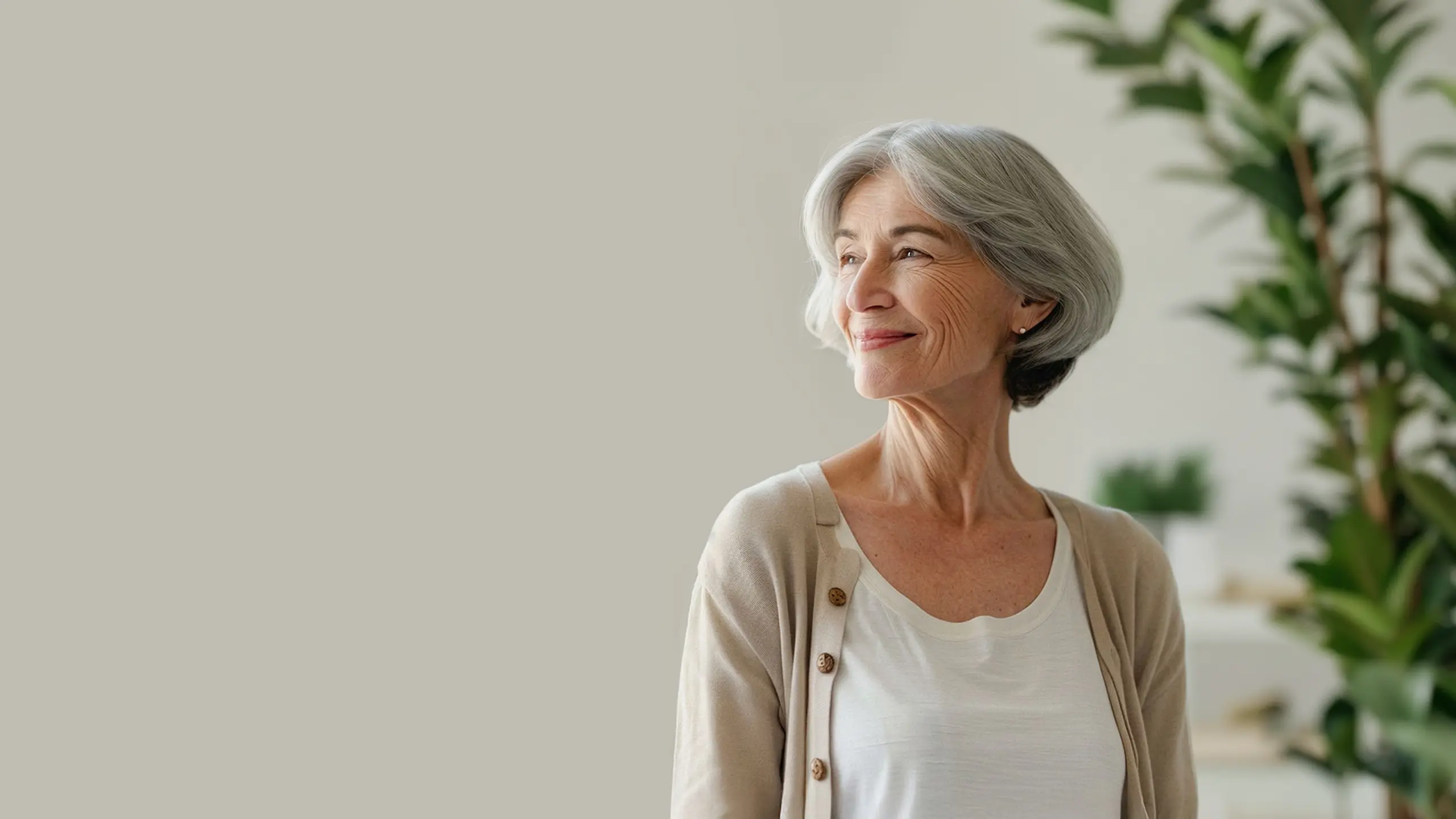 Older woman with gray hair smiles looking out, standing indoors near plants and shelves in soft light.