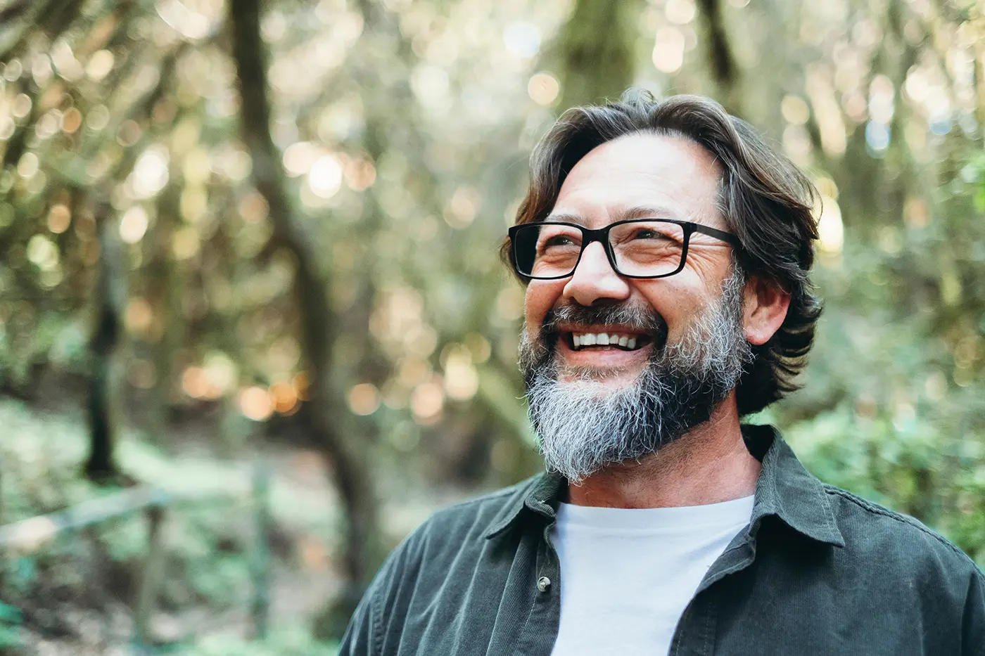 Smiling man with glasses and gray beard standing outdoors with blurred trees in background.
