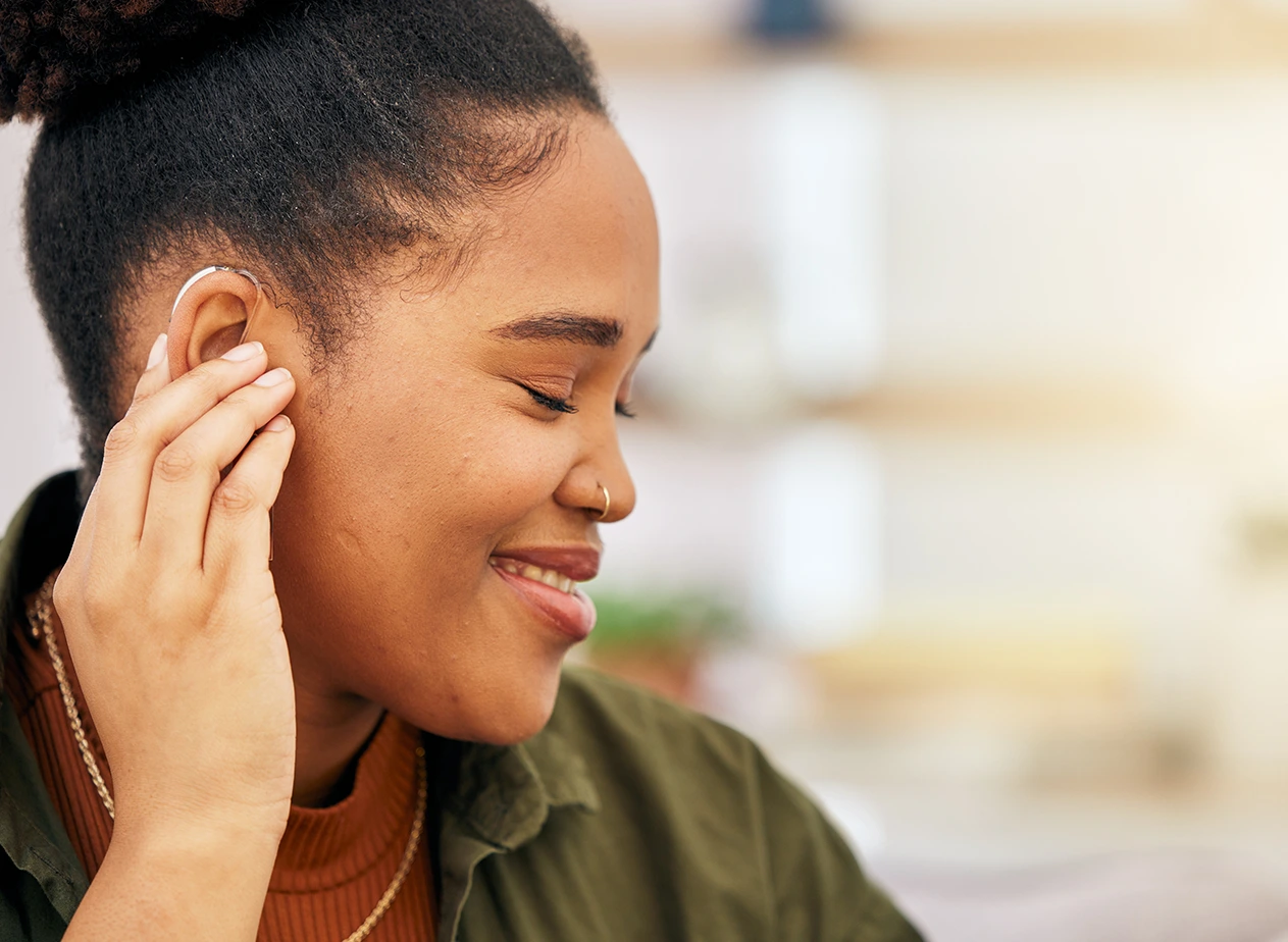 Smiling woman with eyes closed adjusts hearing aid in her right ear indoors.