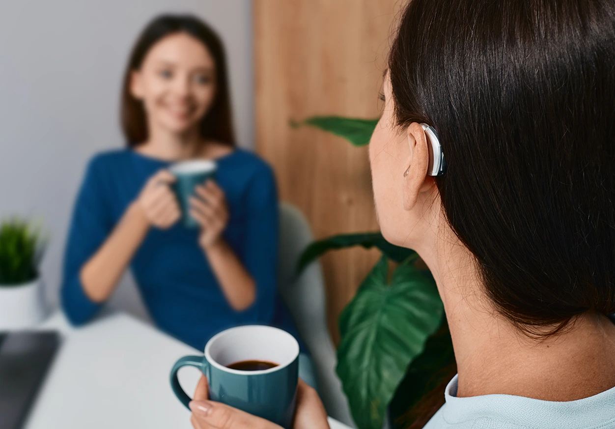 Two women drinking coffee and talking, one wearing a hearing aid, sitting near a plant indoors.