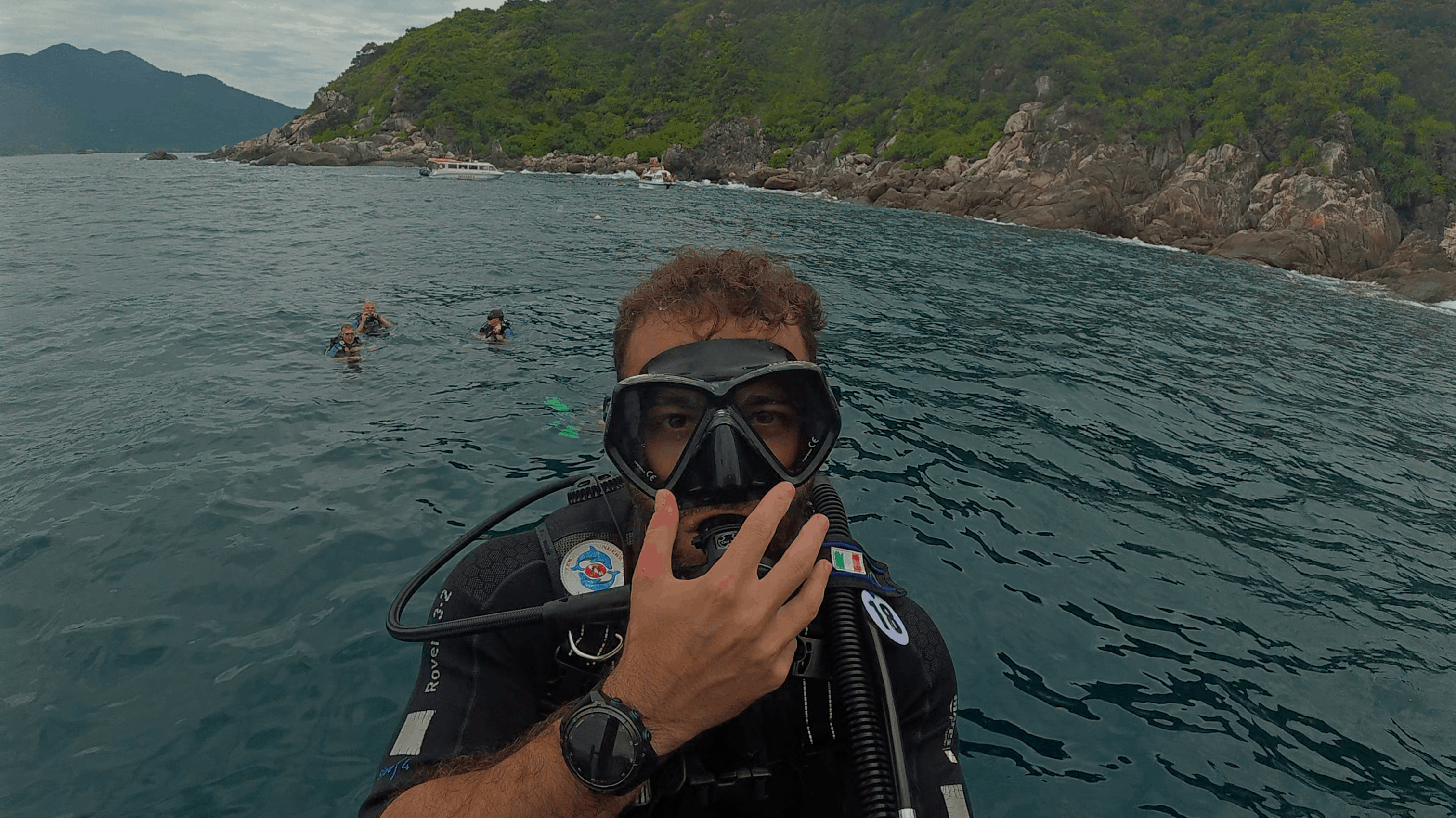 Austin at the surface in mask and gear with divers, speedboats and the island behind at Hon Dai, Cham Islands, Vietnam