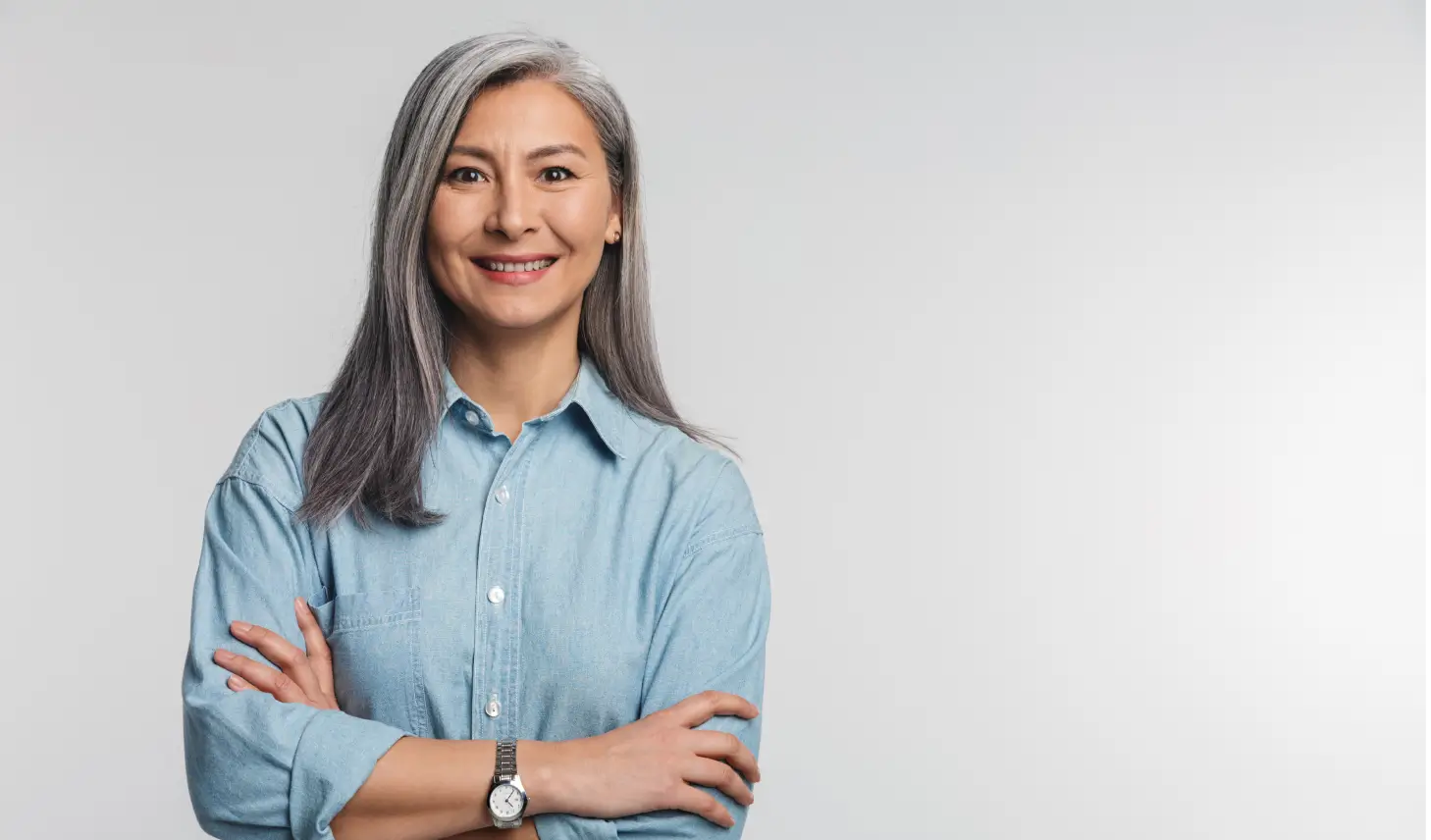 Smiling middle-aged woman with long gray hair, wearing a light blue button-up shirt and watch, standing with arms crossed against a light gray background.