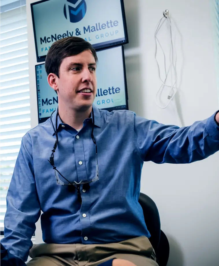 Man in blue shirt with glasses hanging around his neck sitting in front of two screens displaying McNeely & Mallette Family Dental Group logo.