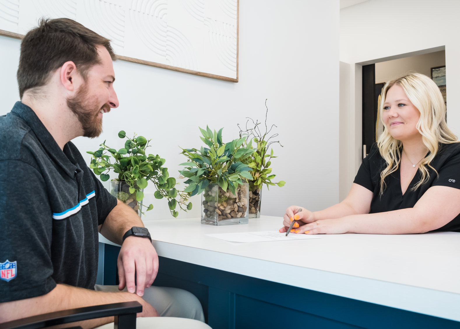 A man and a woman sit across from each other at a desk, the woman holding a pen over a document while they smile.