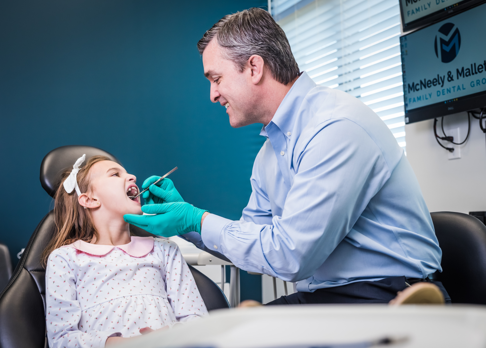 Dentist wearing green gloves examining a young girl's teeth in a dental office.
