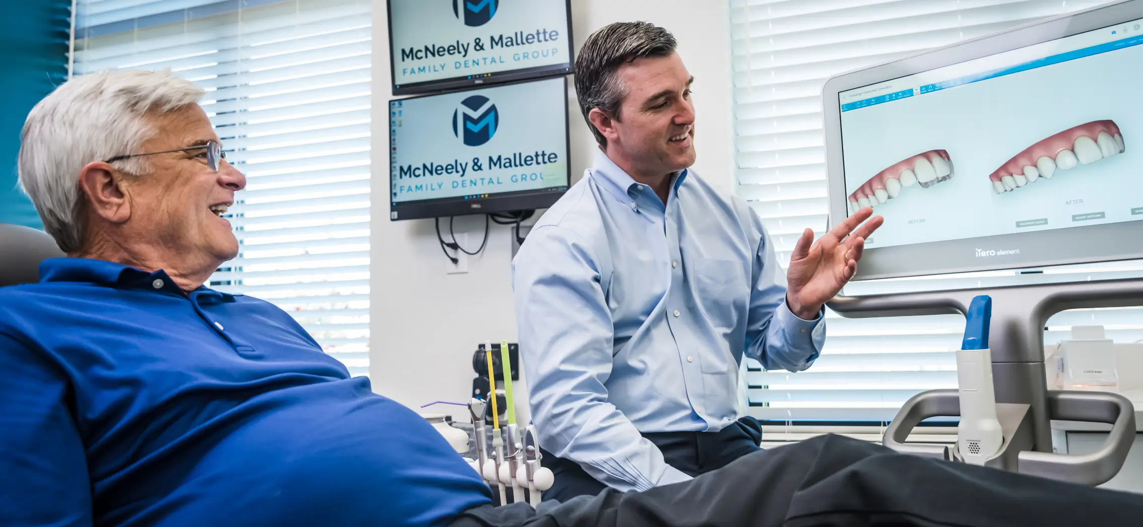 Dentist in light blue shirt explaining dental treatment with before and after images on a screen to an older male patient in a blue shirt.