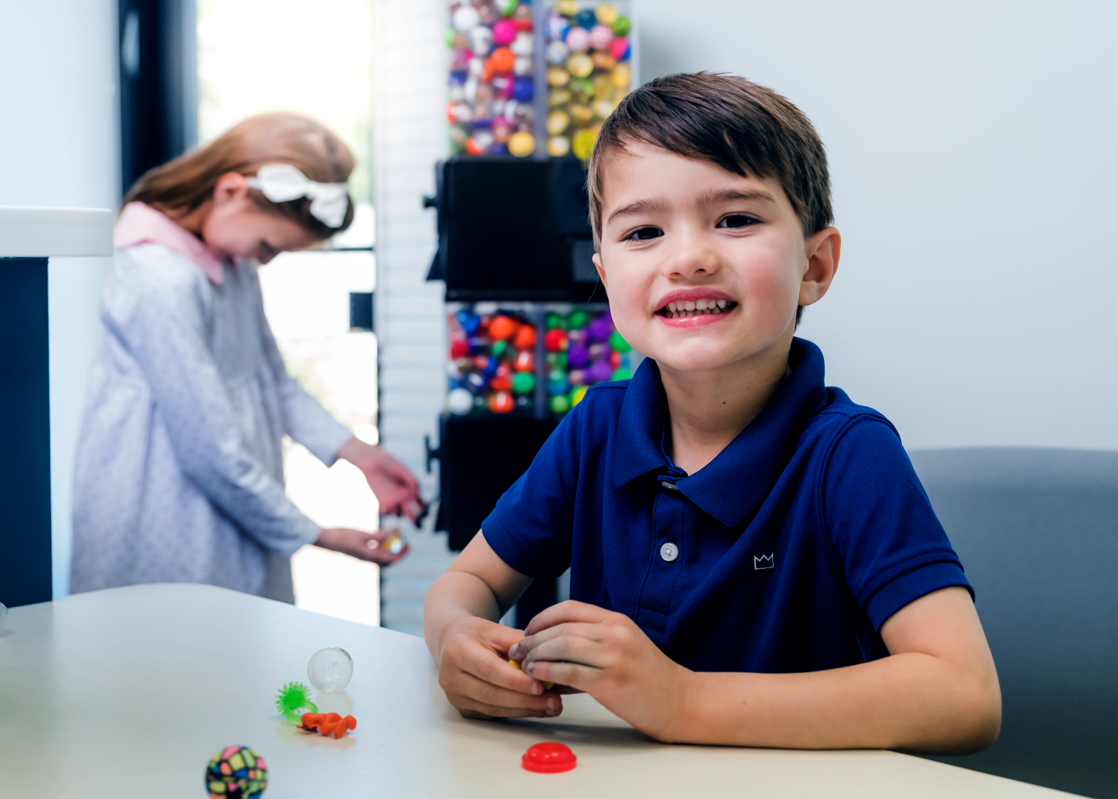 Smiling boy in a navy blue polo shirt sitting at a table with small colorful toys, with a girl in a light dress playing in the background near a gumball machine.