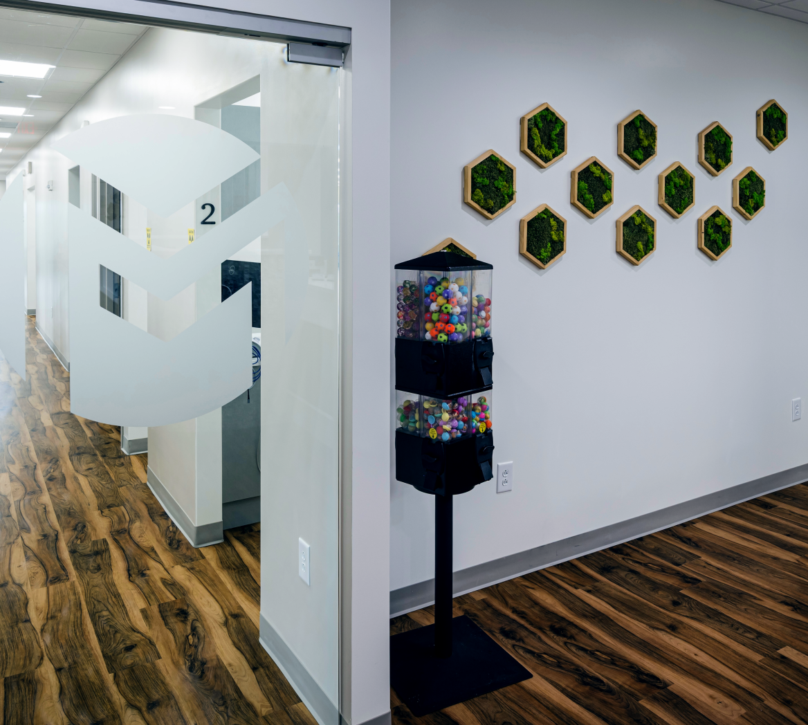 Office hallway with wooden floor, glass door marked with a logo and number 2, wall-mounted hexagonal plant frames, and a black stand holding two gumball machines filled with colorful balls.