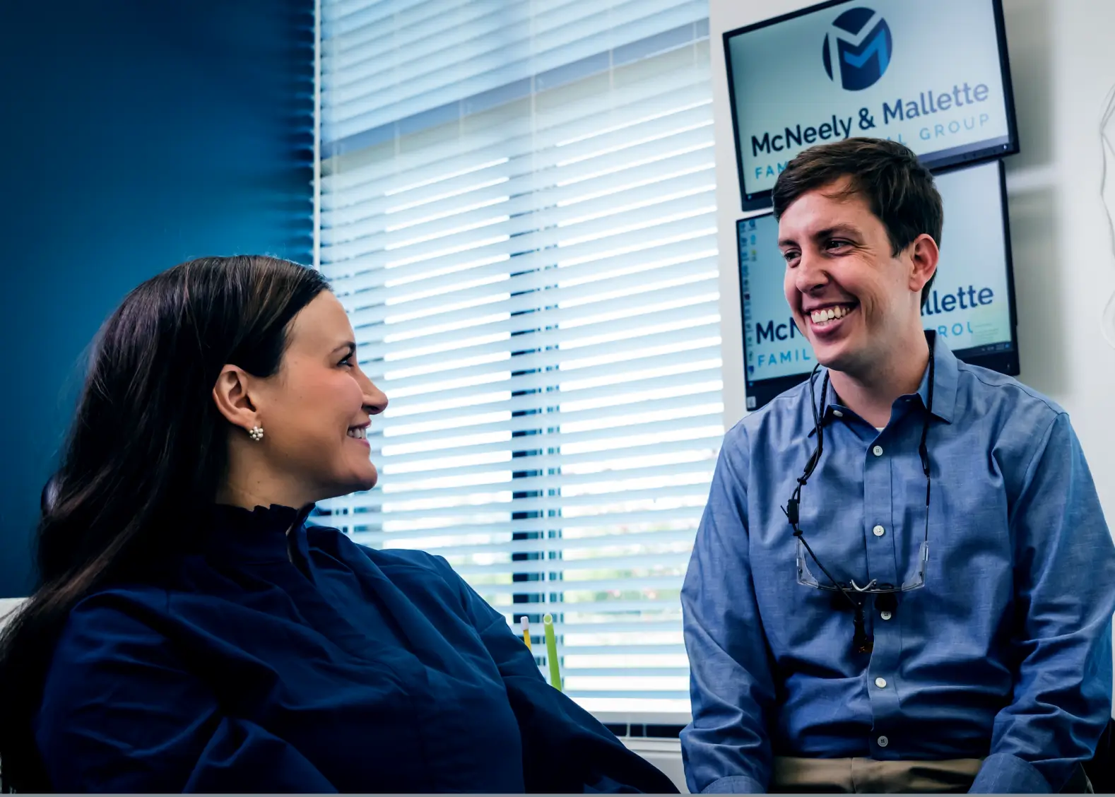 Smiling male dentist talking with female patient in dental office with McNeely & Mallette Family Dental Group sign in background.