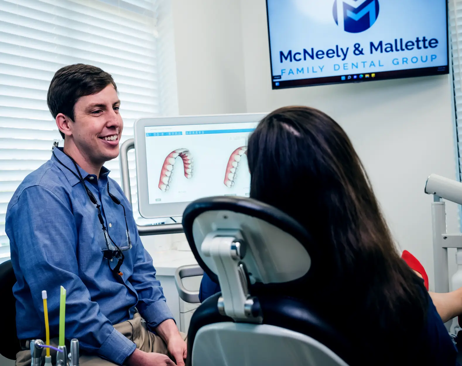 Dentist in blue shirt smiling and explaining dental images on a screen to a seated patient in a dental office.