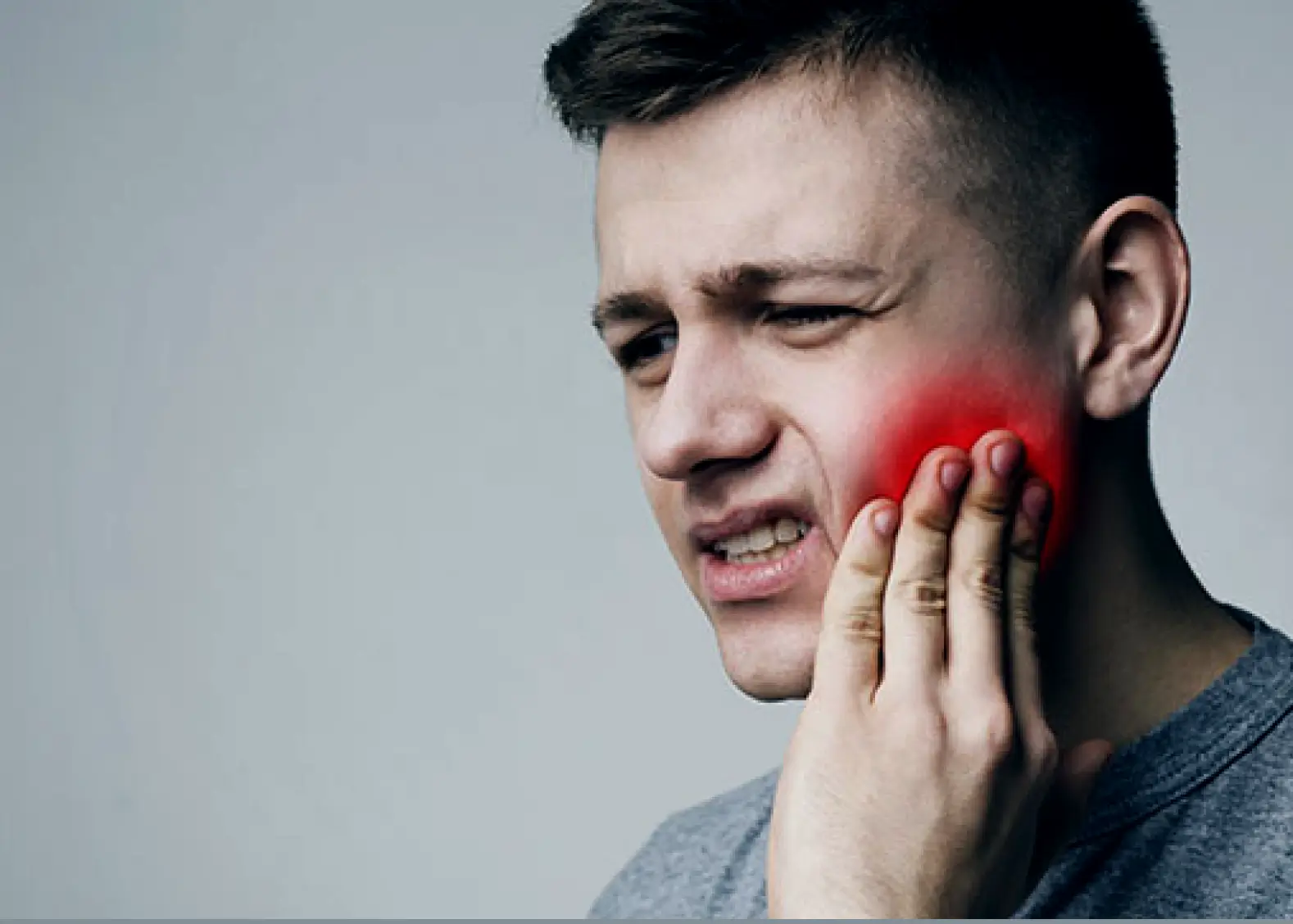 Young man grimacing in pain while holding the right side of his cheek highlighted with a red glow.