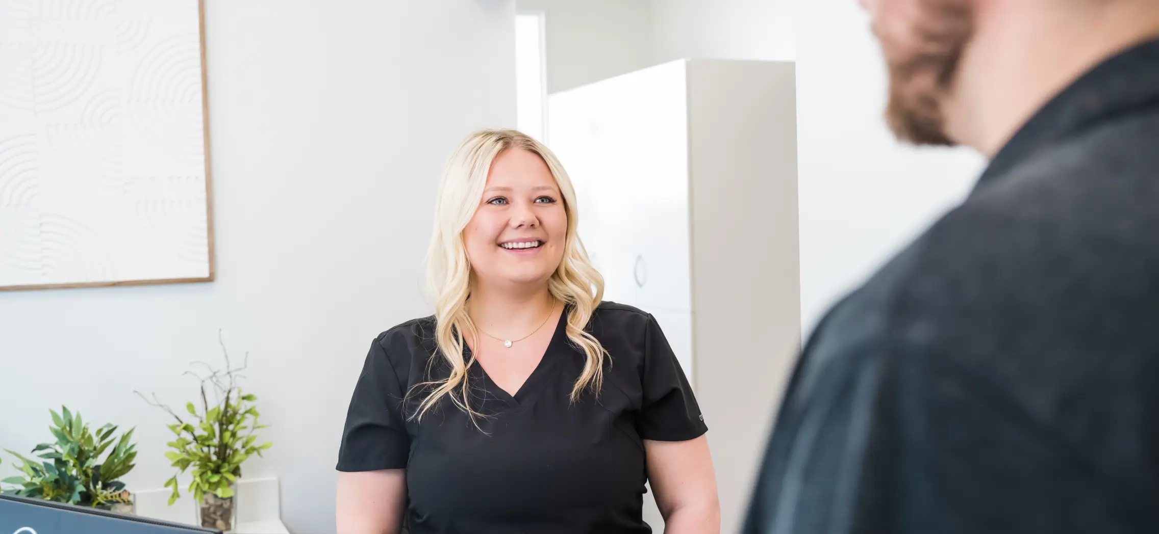 Smiling blonde woman in black scrubs speaking with a man in a bright, modern office.