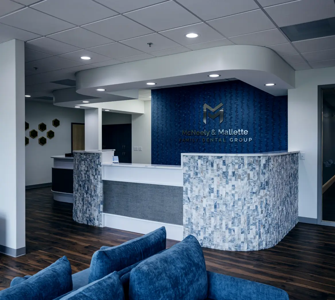 Modern dental office reception area with a blue textured wall displaying McNeely & Mallette Family Dental Group logo, white curved desk, and blue couches.