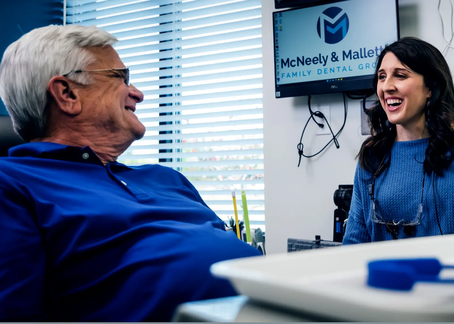 Smiling elderly man speaking with a female dentist in a dental office, with a monitor displaying McNeely & Mallett Family Dental Group logo in the background.