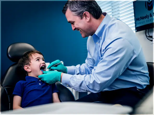 Dentist wearing gloves examines young boy's teeth in dental chair.