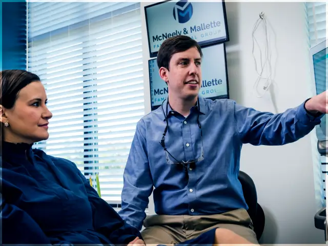 Man in blue shirt pointing at a screen while sitting next to a woman in a dark blue jacket inside an office.