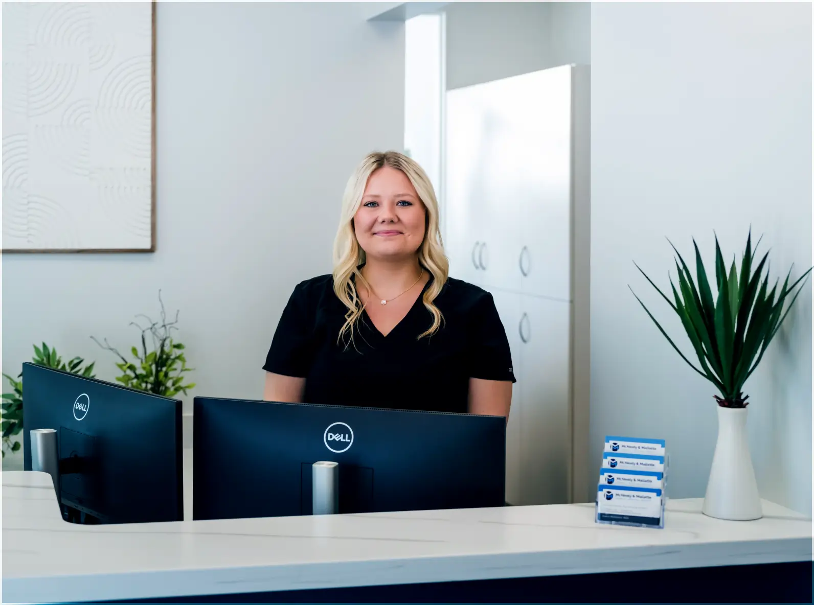 Smiling receptionist with blonde hair sitting behind a desk with two Dell monitors in a modern office.