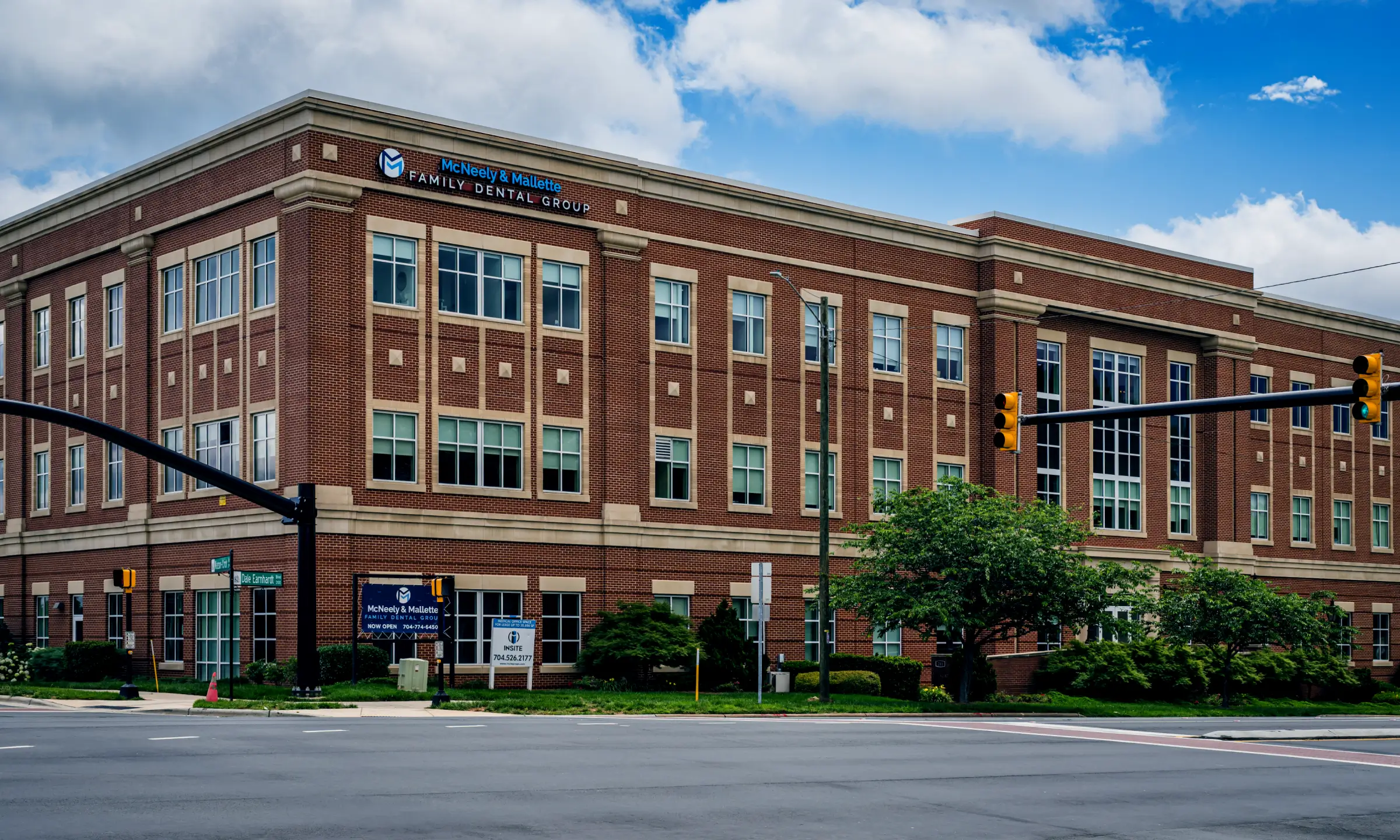 Red brick building named McNeely & Mallette Family Dental Group at a street corner with traffic lights and partly cloudy sky.