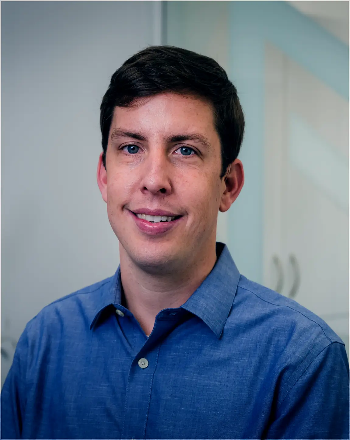 Smiling man with short dark hair wearing a blue button-up shirt in an indoor setting.