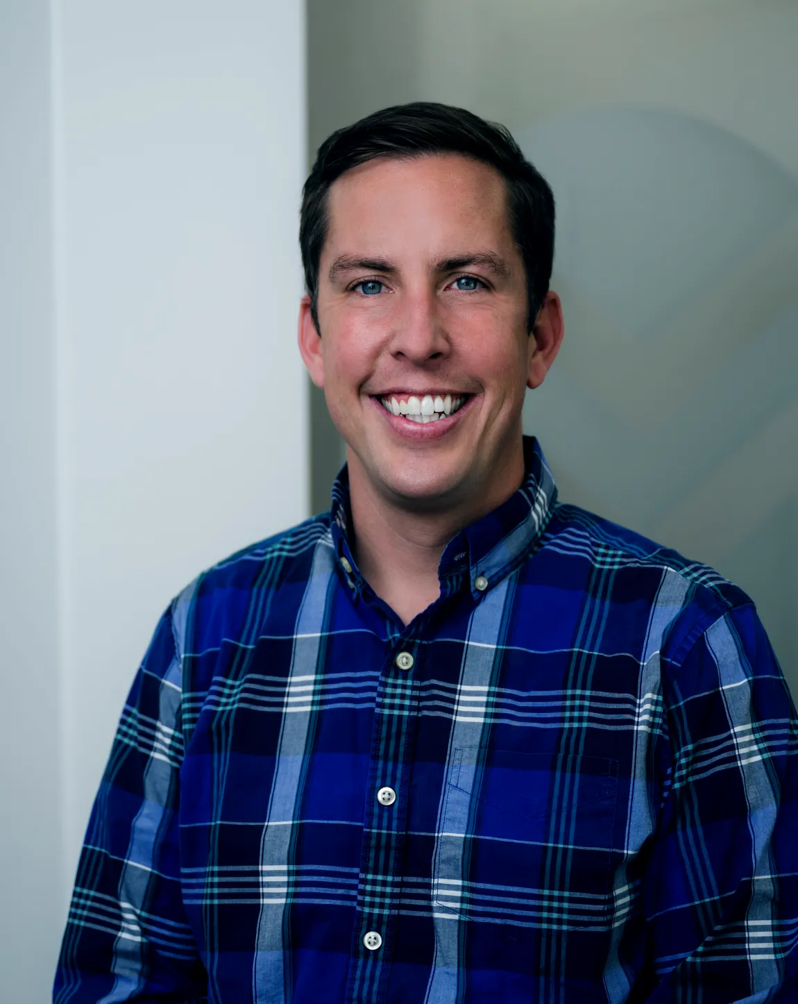 Smiling man with short dark hair wearing a blue button-up shirt in an indoor setting.