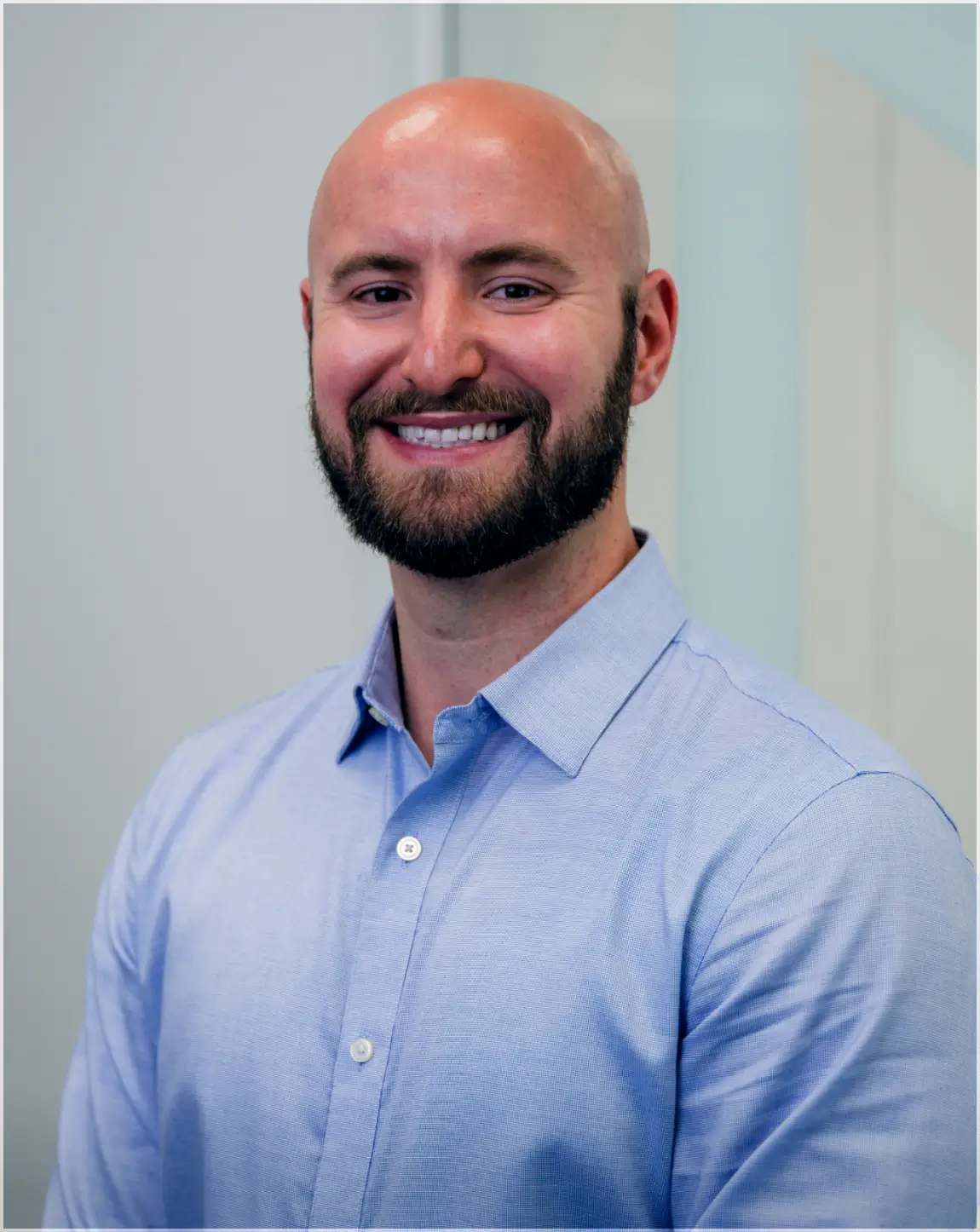 Smiling bald man with a beard wearing a light blue button-up shirt against a neutral background.