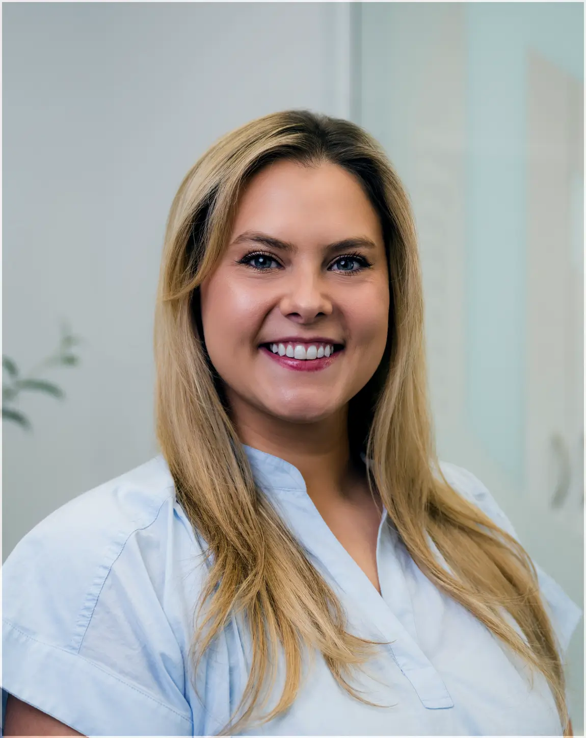 Smiling woman with long blonde hair wearing a light blue shirt against a soft indoor background.