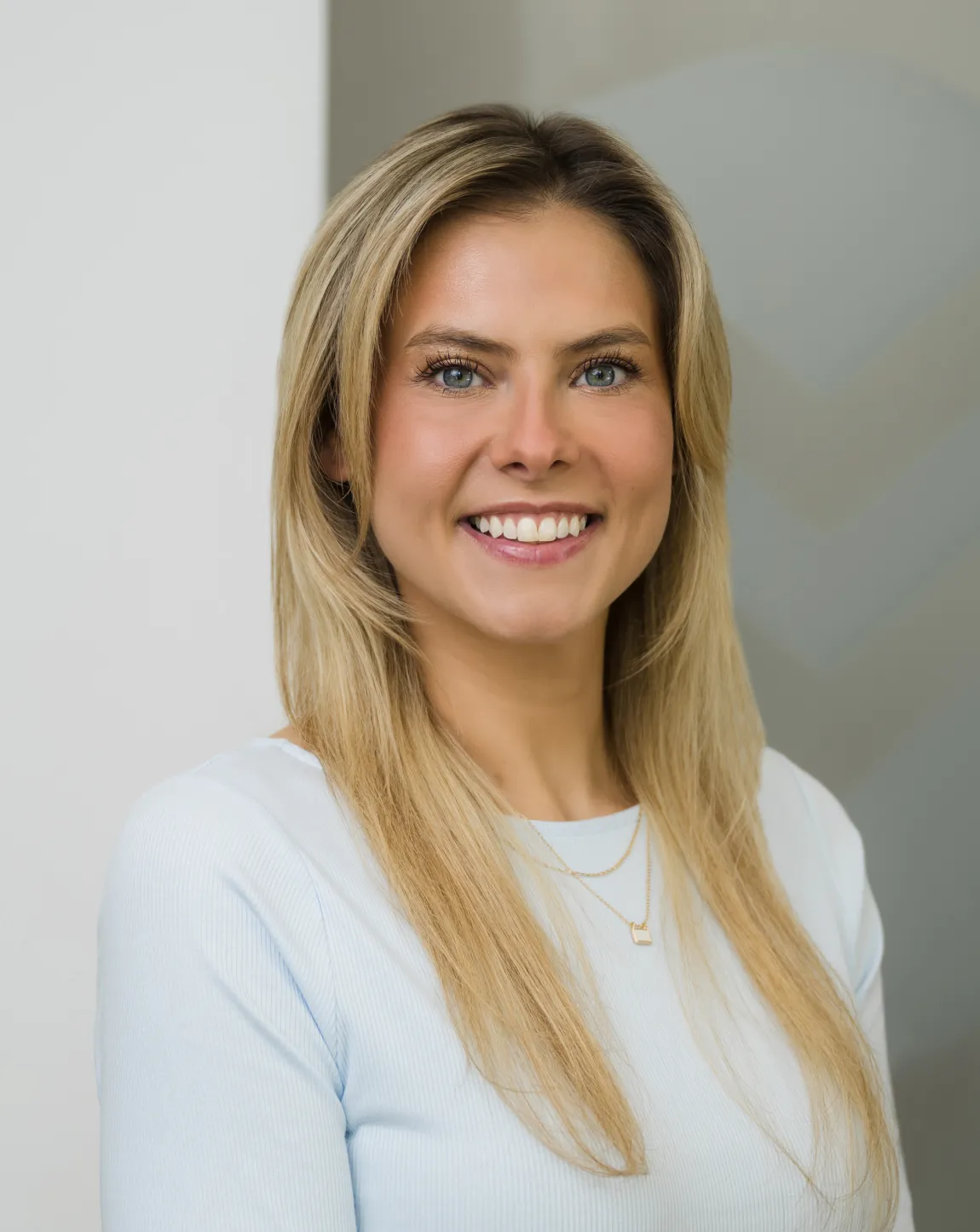Smiling woman with long blonde hair wearing a light blue shirt against a soft indoor background.