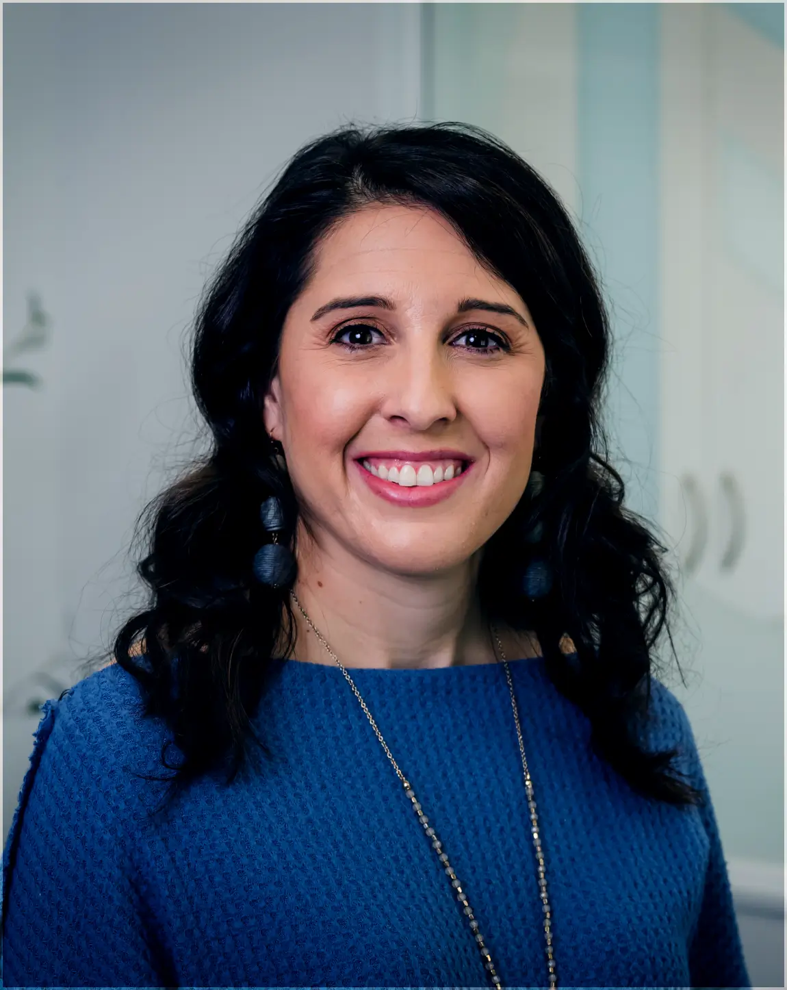 Smiling woman with dark curly hair wearing a blue textured top and a long necklace in an indoor setting.