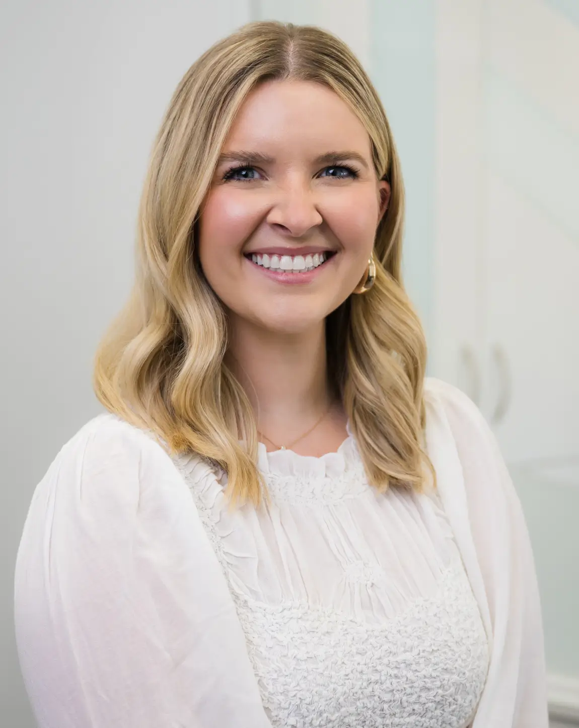 Smiling woman with blonde wavy hair wearing a white textured blouse and gold hoop earrings.